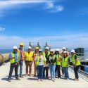 11 people stand on a pier above a body of water. They are wearing safety vests and hard hats. A large pipe is seen on the left.