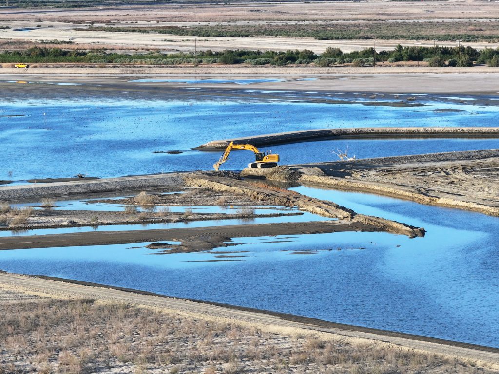 An excavator breaching a berm to allow water to move from the sedimentation basin to the east pond