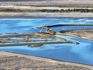 a berm being breached by a yellow excavator to allow water to flow to the other side of the pond